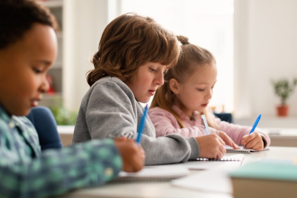 Diverse pupil writing during the lesson at desk in classroom at elementary school, sitting in row
